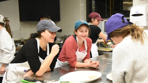 Three students in aprons and baseball caps talking around a metal table.