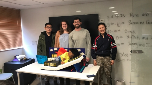 Four students stand in front of their project, which they created out of cardboard and designed to look like a rocket ship. The device supports a doll that represents a small child.