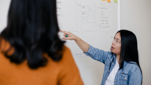 Two women in a classroom with one woman pointing at writing on a board while the other observes