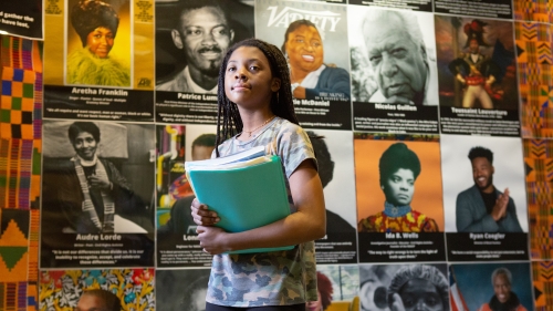 a young Black girl stands in front of a wall displaying prominent Black American Figures