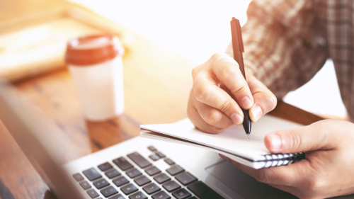 Photo of student taking handwritten notes while learning at their laptop computer.