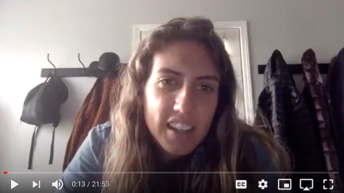 Alexis Niccum in front of a coat rack with coats, a hat, a scarf, and a backpack hanging.