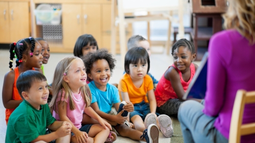 A photo of nine children sitting on the floor of a classroom and watching an adult present a book. The adult has their back to the camera and the text of the book is not visible.