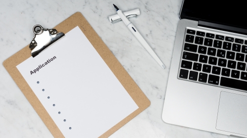 A clip board with a sheet of paper titled "Application" next to a white pen and laptop to the far right