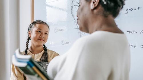 A photo of a girl with braids and an adult teacher standing in front of a dry erase board and talking.
