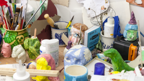 A snapshot of a ceramicist's studio. Paintbrushes and glue and ceramics all over a table. 