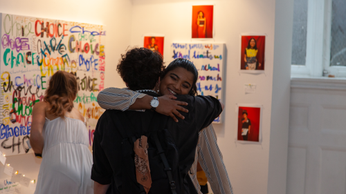 Visual Arts Administration students embrace at an exhibition. Two students hug in the foreground; one is facing the camera with a smile on her face. In the background, another student adjusts a painting installed on the wall.