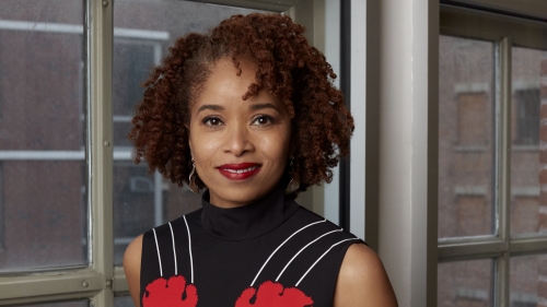 Dr. Nicole R. Fleetwood stands next to a window wearing a black dress with white stripes and red flowers.
