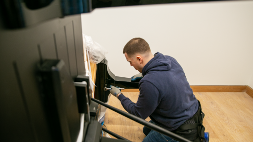 A worker in a blue sweat shirt installs a leg on a Steinway piano that is resting on its side.