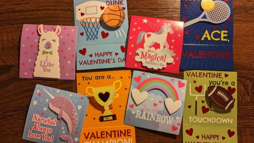 A pile of Valentine's Day cards on a table. Half are decorated with smiling animals, rainbows, and hearts; the other half are sports-themed.