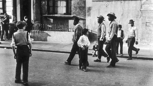 A street scene in post-war Britain showing a white adolescent watching some black men walk down the street