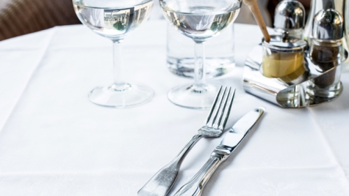 Image feature round dinner table covered with fancy white linen, shiny cutlery, and two water glasses filled halfway