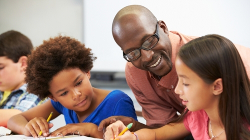 Educator assists two elementary school students as they sit at their desk completing class assignment. An Blac female is seated to the left of the teacher, while a Latina students sits to the right of the male teacher wearing Black frame eye glasses.