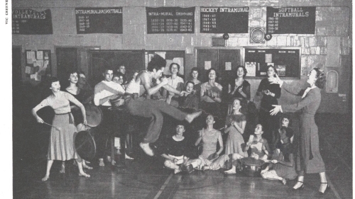 A black-and-white 1951 photo of a group of dancers in a a gym. Some women are holding drums while a man jumps energetically in the center. The caption reads "Martha Hill Directs a lively modern dance session at NYU."