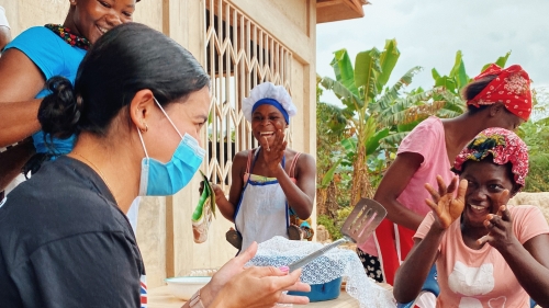 A student wearing a surgical mask smiles and claps with a group of Ghanaian women