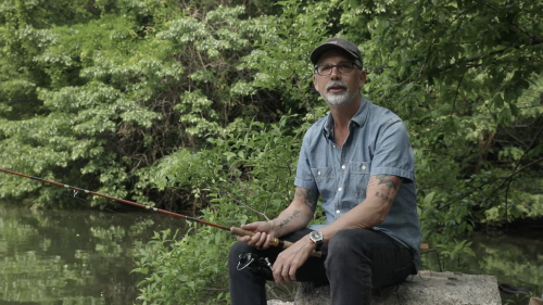 Stephen Duncombe sits on a rock in Central Park holding a fishing rod. 