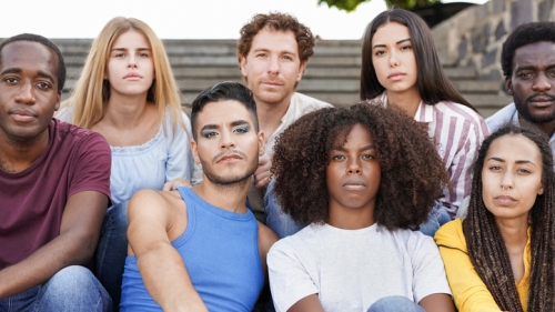 Image features a photo of 8 Queer and Trans Youth. The students are sitting outside on concrete staircase., in a grouping that finds 3 of the students sitting in front of second grouping, of 5 students sitting behind the. The students are looking straight forward at the camera with assertive , and independent expressions.