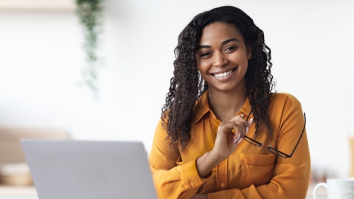 Image captures student sitting at a desk, holding her eyeglasses and glancing at her laptop screen.