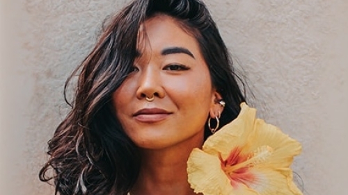 Liz has black wavy hair and wears a garland of large hibiscus flowers 