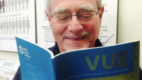Image captures headshot of Norm Fruchter reading a copy of Voices of Urban Education. Norm wears eye glasses and smiles as he reads the open-access journal.