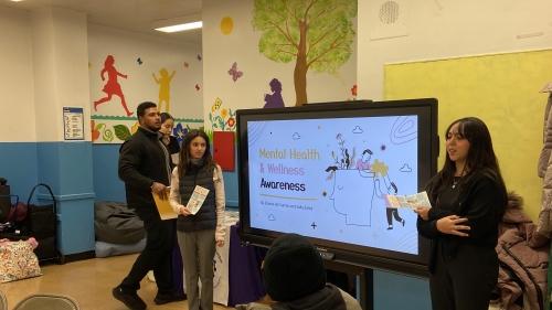 Two students presenting to a classroom, standing next to a monitor that reads "Mental Health Wellness & Awareness"