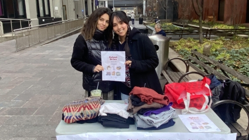 Two students standing at a collection table outside at NYU holding a flyer that says "School Drive"
