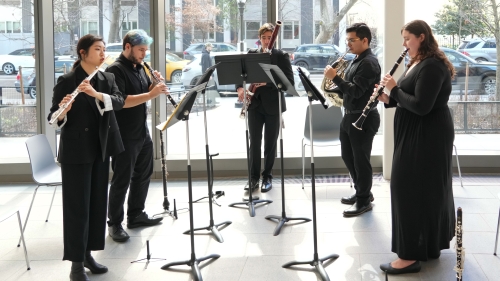 Five brass and woodwind performers playing in front of large sunny windows