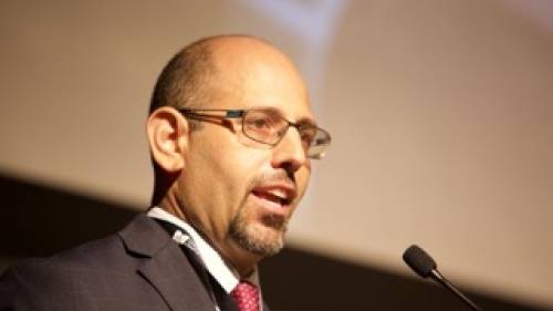 Headshot of Josh Thomases. Photo features man speaking into a microphone attached to a podium, as  he wears a red tie and glasses.