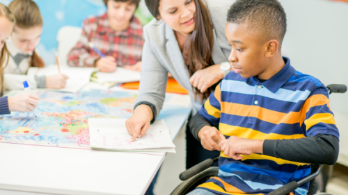 Student in wheel chair sitting at desk with teacher pointing at paper