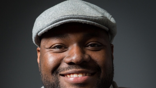Headshot of Tim Ojetunde. The photo features a man in front a dark gray background, as he smiles at the camera wearing a gray hat.