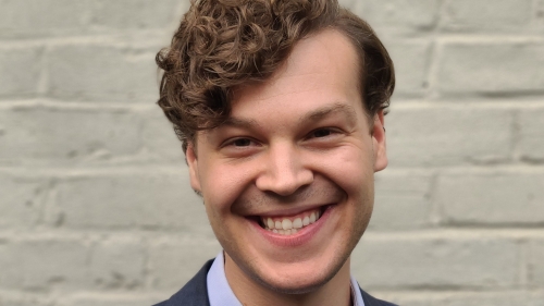 Image captures a headshot of Trevor Baisden. Photo features a man smiling at camera as he stands in front of a brick wall, light blue shirt, and navy blue sportcoat