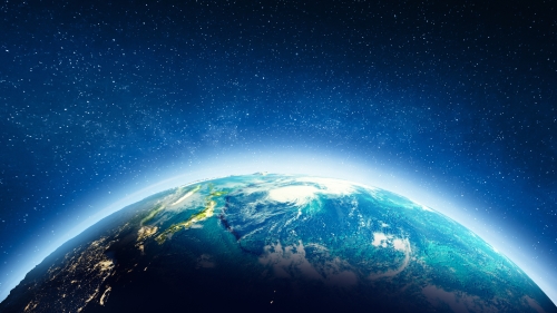 Image captures a photo of the planet earth from space. The horizontal photo displays a quarter of the globe, outlined with a white sunlight against the backdrop of the darkness of space. At this distance, the picture of the earth could be dark blue marble, covered with patches of green vegetation, and white clouds and atmosphere. This zenith image of the earth is in focus and reflects the sunlight into the otherwise dark space vacuum.