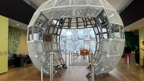 Photo of a food studies art exhibit with a sphere structure up front, with two signs above reading "EATING" and "TRADING"