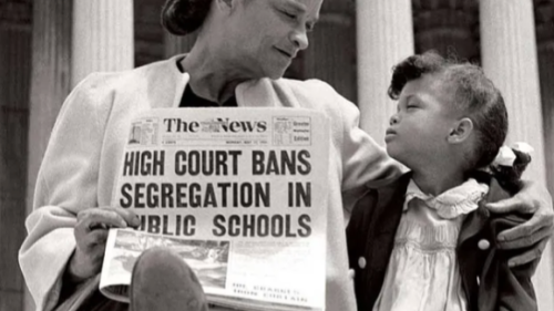 Image captures a mother and daughter sitting on the step of the US Supreme Court. The two look deeply in each others eyes, as the mother holds a newspaper in her hands. The newspaper headline reads, "High Court Bans Segregation In Public Schools". Behind the mother and daughter, one can see the columns of the Supreme Court.