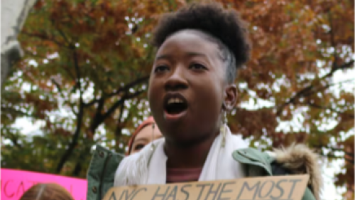 Image captures a student protesting for an end to segregation in NYC schools. This high school student holds a protest sign in front of her which reads, NYC has the most segregated school system in America. We need equity and integration. The student is wearing a green coat and white scarf.