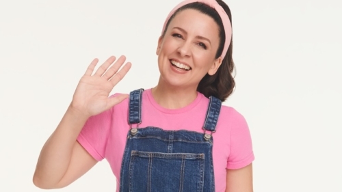Ms. Rachel smiles and waves; she wears a pink shirt and blue overalls.