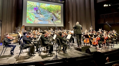 Full orchestra performing on stage in front of screen with images