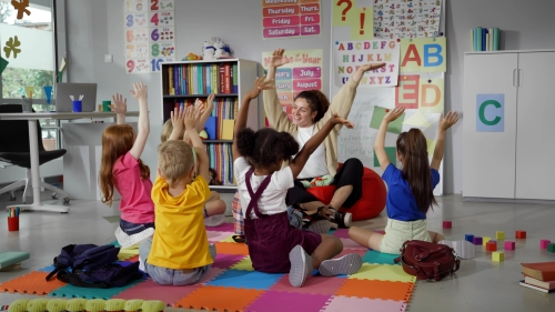 Elementary school teacher sit in class on floor with little pupils and moving hands up.