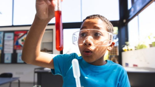 Image captures young junior high school students working on a STEM experiment in a classroom lab. The student, wearing a blue shirt, is examing the contents of a vile of liquid. The student also wears security glasses to protect his eyes.  