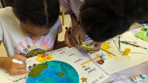 Image captures young student artists at work inside of a New York City classroom. This STEM assignment finds two students at their desk, coloring in a water of globe, as replicated on the back of their paper plate model. One student wearing a white colored t-shirt, and fully seated, holds a marker in her hand as she surverys her work. Another student wearing a light purple colored top looks to afix a yellow item to their collaborative assignment.