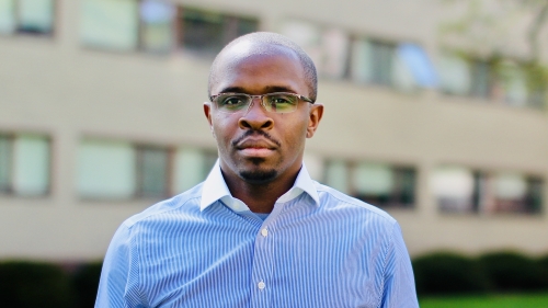 A black man wearing a blue oxford shirt and glasses looks at the camera