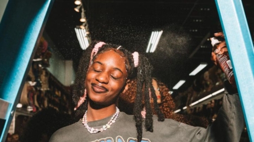 Image captures a performance shot of student artist Divinity-Nix Sow. This student versed for her lyical and poetry skills, wears a oversized black shirt referencing the music group Outkast. She is wearing hair up with two side ponytails on each side. Divinity is facing a lighting source, and holding a can of hair spray.