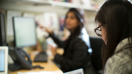 A student looking at a computer. Behind her, out of focus, is another person gesturing toward a computer screen.