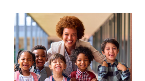 Image captures a teacher leaning in to embrace the five elementary school students with her. Both teacher and students are standing in their schools's outer hallway.. The five littles children each wear backpacks and smile as the photo is taken.