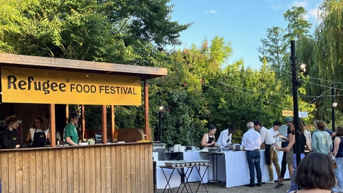 People serve food outside a stand that reads, "Refugee Food Festival"
