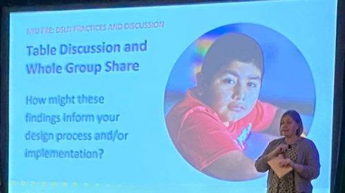 Image caputresa photo of NYU Metro Center PRE Researcher, Sara McCallister, standing before the display of her presentation slides. The image centers around a presentation slide with the image of a middle school child wearing a red shirt. Researcher Sarah McCallister stands on the dais holding note cards in her hands. 