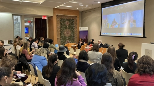 A crowd of people listens to a panel; 3 of the panel speakers sit in front, while 1 panelist is projected onscreen