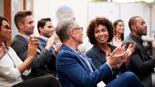 Image captures the a seated conference audience. The photo centers around two audiences members sitting together.. Nearest the camera lens, is a man wearing a blue suit, as he applauds. Next to him, is Black Woman who is in conversation with the man in the blue suit.