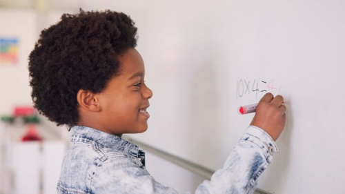 Image captures a child working about a problem on the whiteboard in his classroom. This elementary school aged child writes using a red marker on a whiteboard. The child wears an blue, acid-wash colored shirt and a short afro.