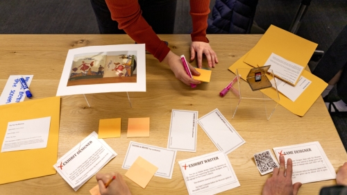 Students hands are shown working around a table with papers outlining their roles and standing exhibits including a Super Smash Bros cartridge